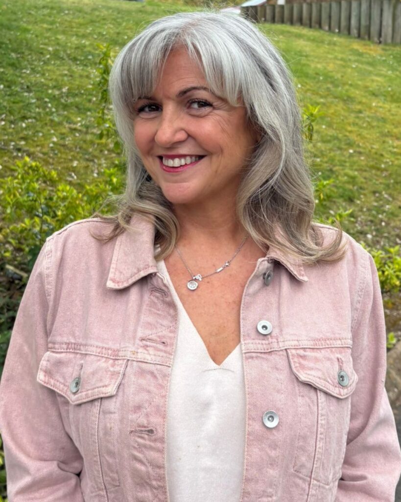 A person wearing a pink denim jacket over a white top smiles at the camera. They are standing outdoors on a paved path with a grassy slope in the background.