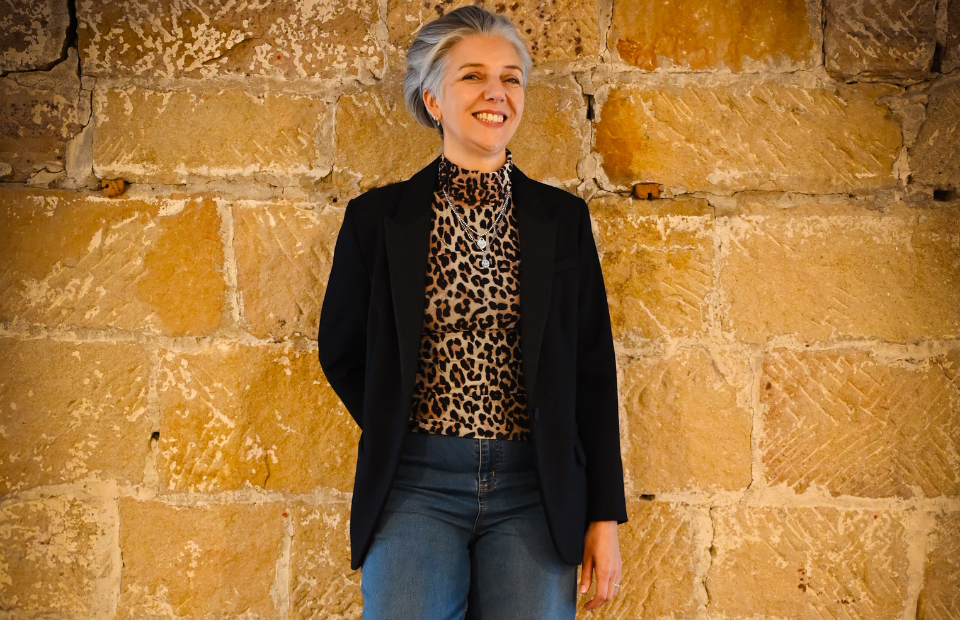 A person in a black blazer and leopard-print shirt stands against a stone wall.