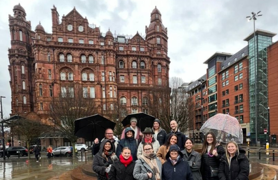 A group of people stand in front of a large red brick building on a cloudy day. Some people are holding umbrellas.