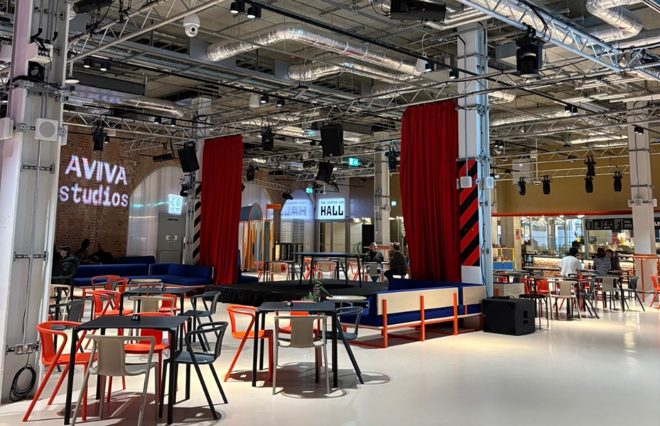 A spacious indoor venue with seating areas, industrial looking pipe work on the high ceiling, a stage, and "AVIVA studios" signage on the wall.