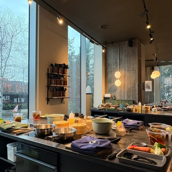 A cooking class setup with multiple workstations, each equipped with cooking pans, bowls, utensils, and ingredients like carrots and celery. Large windows provide natural light and a view of trees and buildings outside.