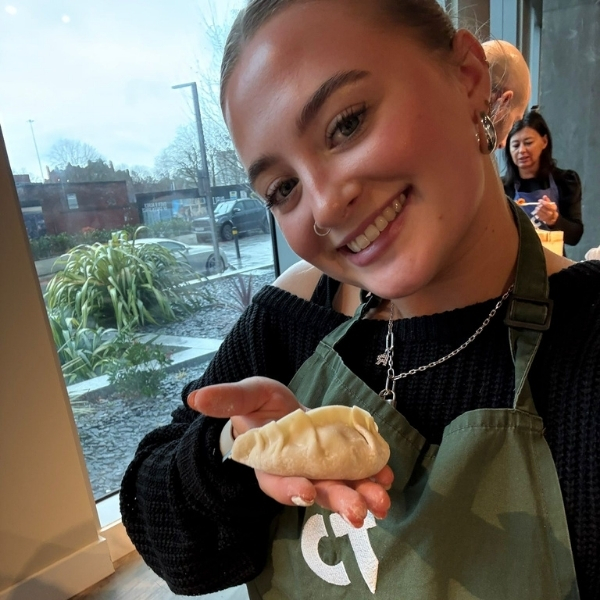 A person smiles looking at the camera, while holding a freshly made gyoza dumpling in their hand. They are wearing a green apron and a black sweater.