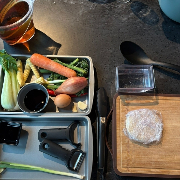 A collection of ingredients for cooking, including vegetables like baby corn, carrots, and broccoli, along with an egg, shallots, garlic, chili, and soy sauce. Tools like peelers, tongs, and a cutting board are also visible.