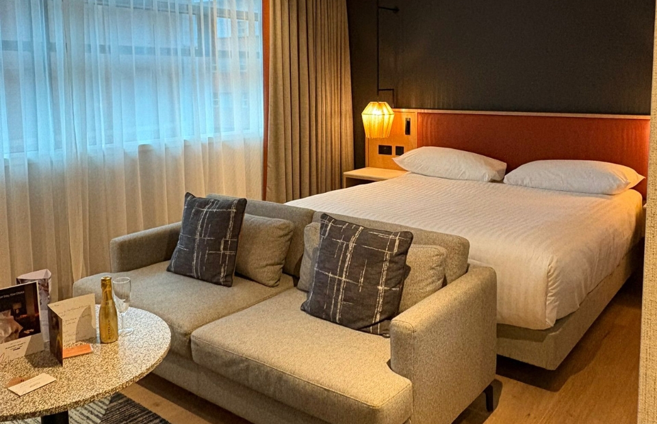A hotel room with a sofa, a bed, and a bedside table with a lamp.