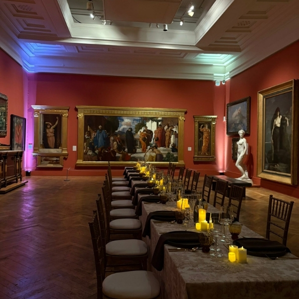 A long dining table is set with place settings and lit candles in a gallery room with paintings on the walls.