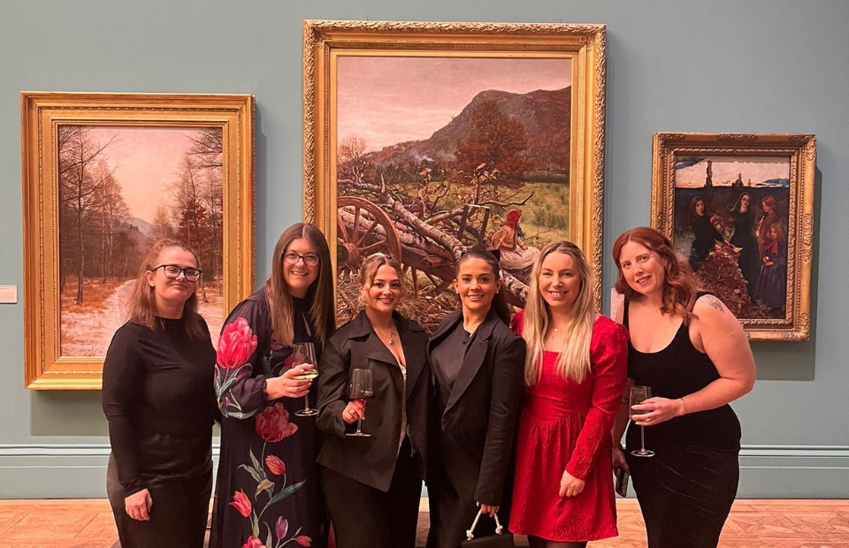 A group of six people stand in front of three large gallery paintings with gold coloured frames.