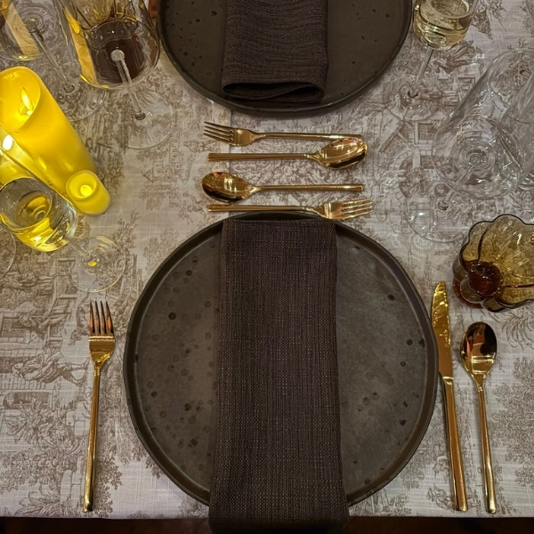 A table setting with dark plates, gold cutlery, dark napkins, and lit yellow candles on a patterned tablecloth.