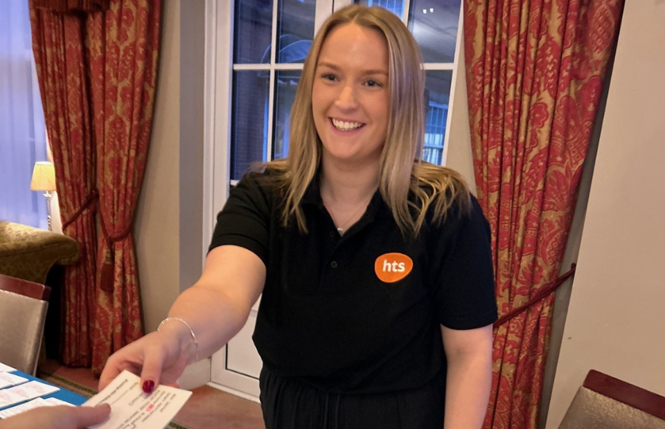 A person in chard of event delivery wears a black polo shirt with an orange 'HTS' logo. She hands a white card to another person, with ornate red curtains in the background.