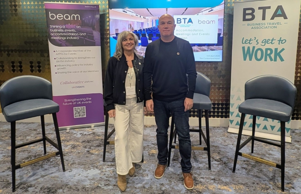 Two people stand between two tall chairs in front of banners that read "beam" and "BTA - BUSINESS TRAVEL ASSOCIATION Let's get to WORK".