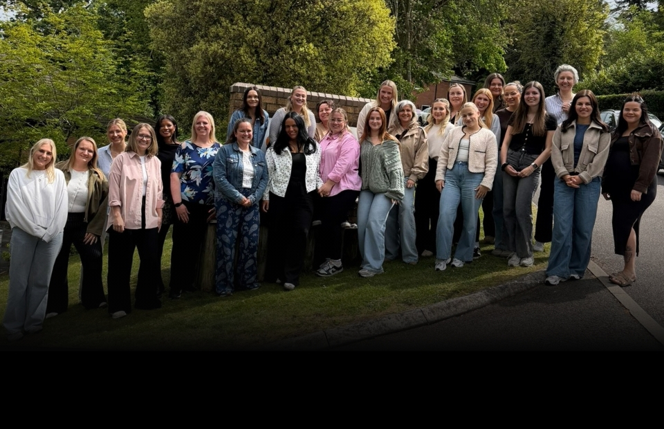 A group of people pose for a photo outdoors on a sunny day, with trees and foliage in the background.