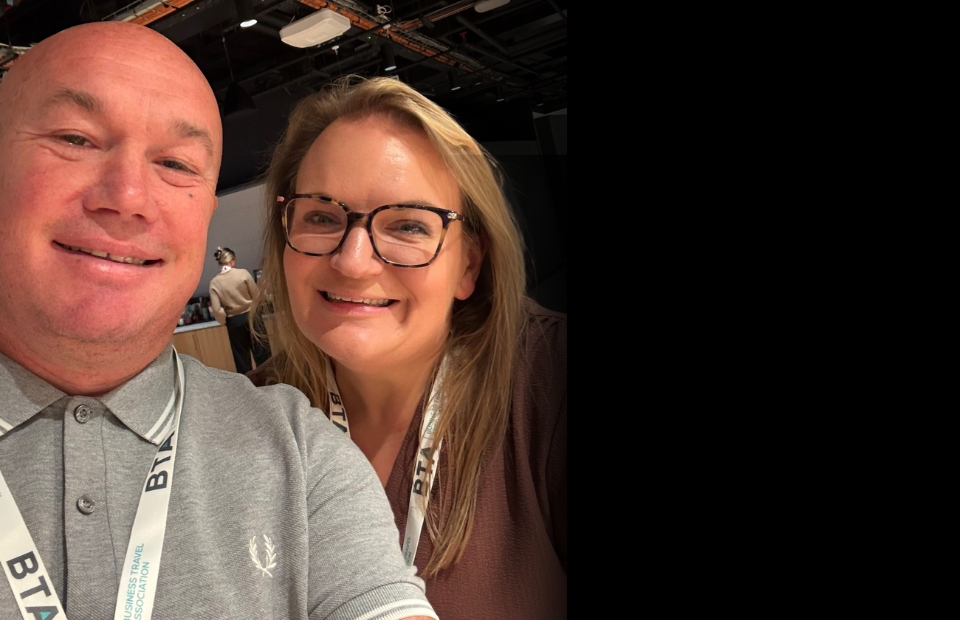 Two individuals wearing lanyards from the Business Travel Association pose for a selfie in an indoor setting.