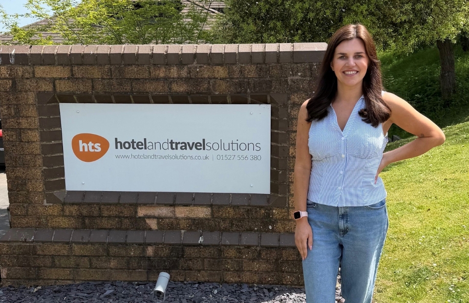A woman stands in front of a brick sign that reads "HTS Hotel and Travel Solutions" with "www.hotelandtravelsolutions.co.uk | 01527 556 380" below it. The sign is set against a background of trees and a blue sky.
