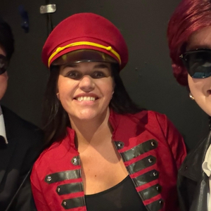 A woman in a red military-style jacket and matching hat smiles at the camera, flanked by two other individuals wearing sunglasses.