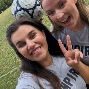 Two women in grey t-shirts with the word "UMPIRE" printed on them take a selfie on a grassy field next to a soccer ball. One woman holds the ball and smiles, while the other makes a peace sign with her fingers.