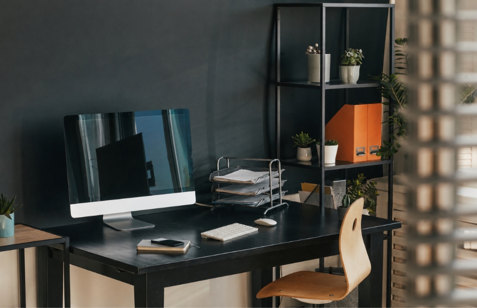 A modern home office setup with a computer, keyboard, mouse, and document tray on a black desk, next to a black shelving unit filled with plants and office supplies.
